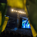 Arquibancada iluminada à noite com bandeiras verde-amarelas e alto-falantes do estádio ao fundo.