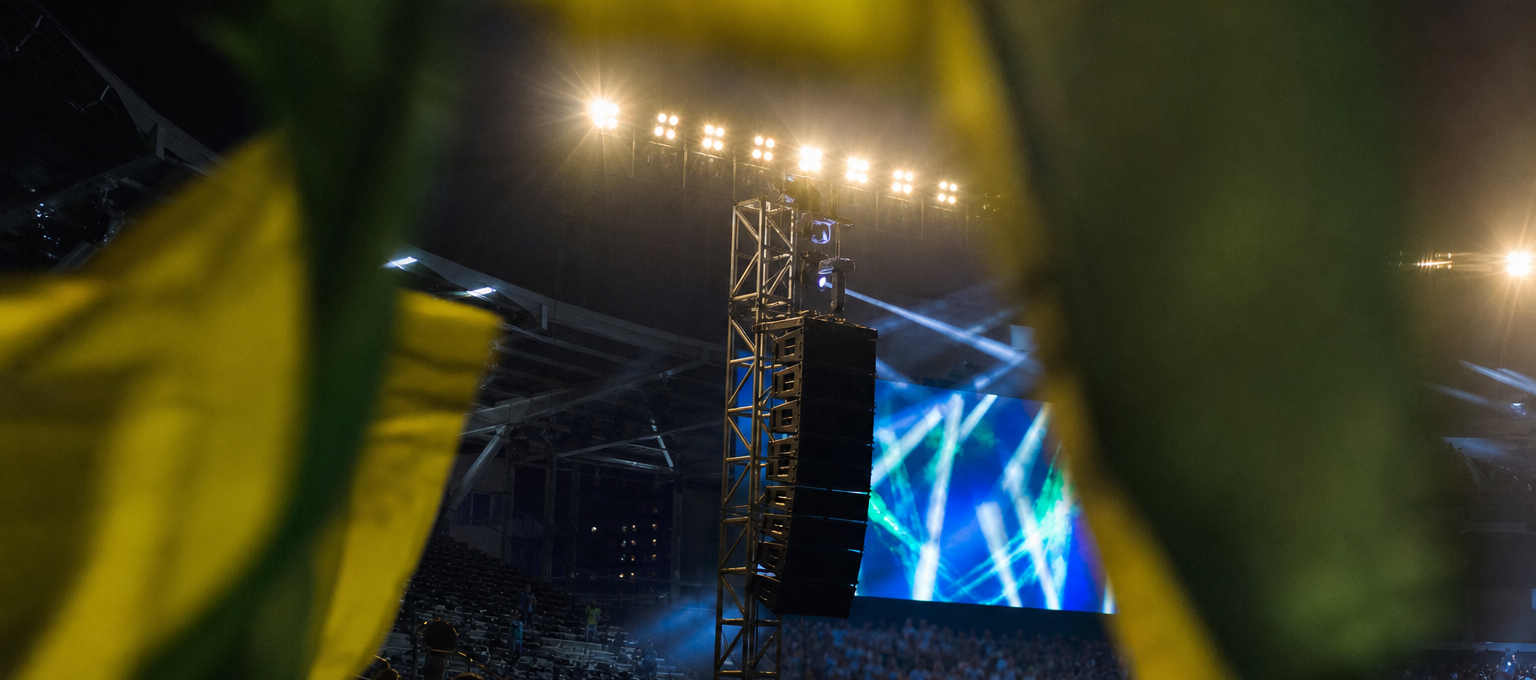 Arquibancada iluminada à noite com bandeiras verde-amarelas e alto-falantes do estádio ao fundo.