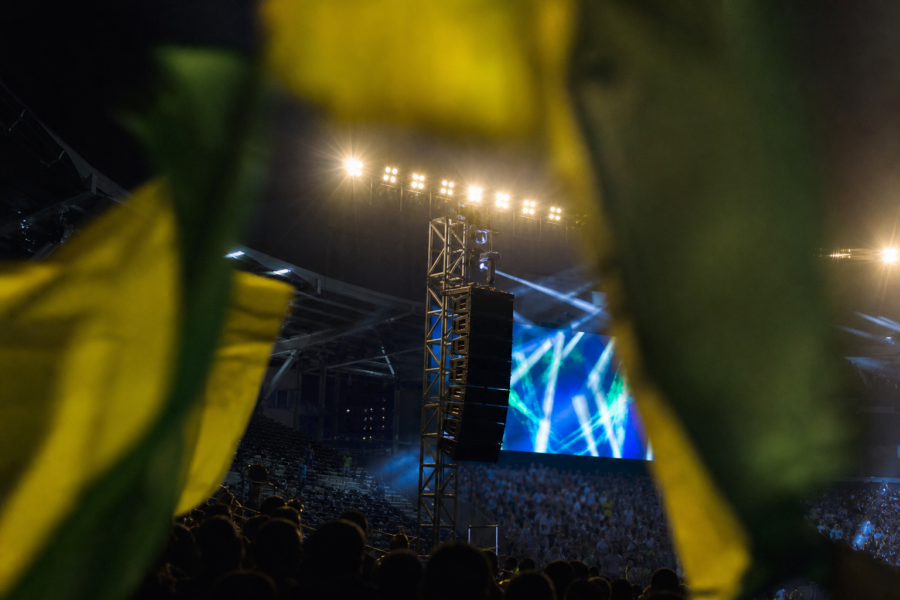 Arquibancada iluminada à noite com bandeiras verde-amarelas e alto-falantes do estádio ao fundo.