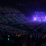 Arena de show com milhares de lightsticks acesos e palco iluminado ao fundo.