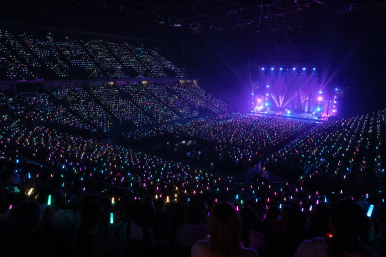 Arena de show com milhares de lightsticks acesos e palco iluminado ao fundo.