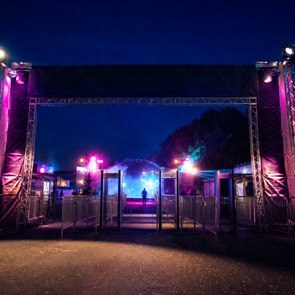 Entrada iluminada de um grande festival à noite, com torres de luz e público em silhueta distante