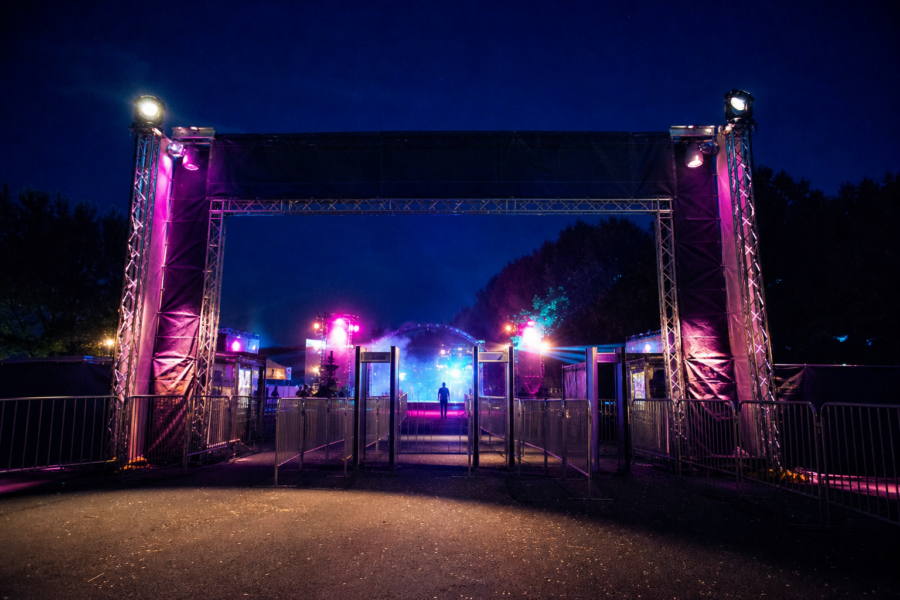 Entrada iluminada de um grande festival à noite, com torres de luz e público em silhueta distante