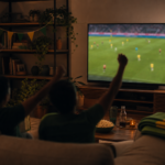 Sala de estar com TV gigante transmitindo futebol e torcida vista de costas