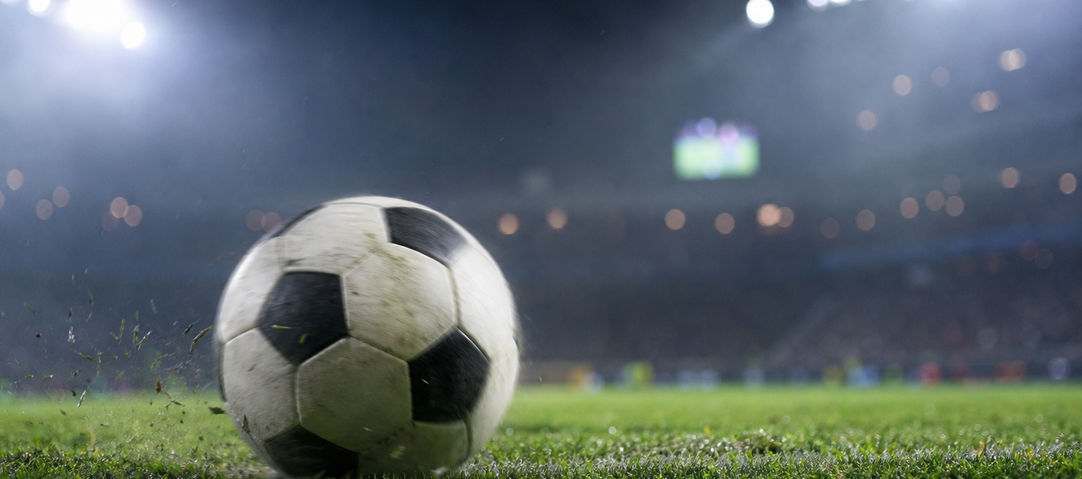 Bola de futebol genérica no gramado com estádio iluminado ao fundo, telão desfocado e clima de jogo.