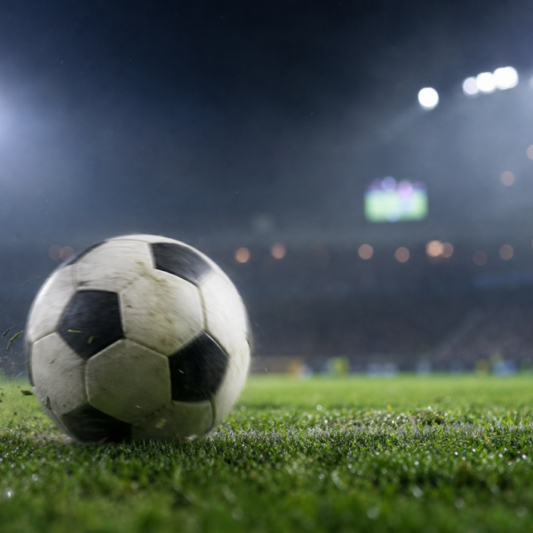 Bola de futebol genérica no gramado com estádio iluminado ao fundo, telão desfocado e clima de jogo.