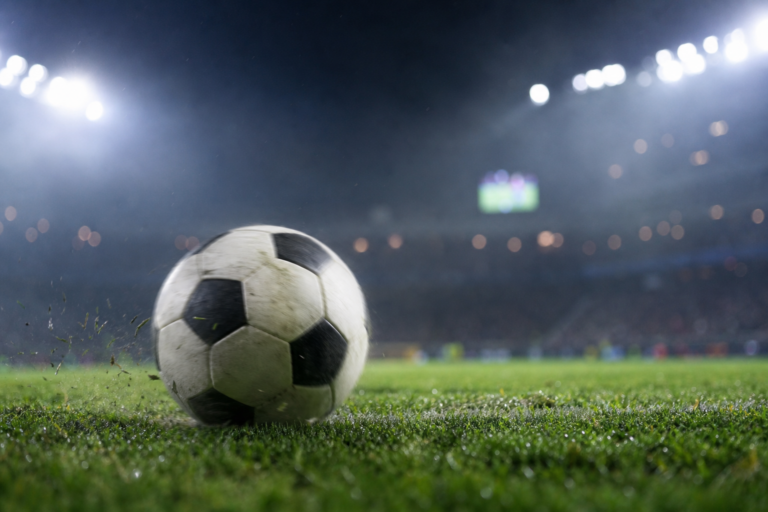 Bola de futebol genérica no gramado com estádio iluminado ao fundo, telão desfocado e clima de jogo.