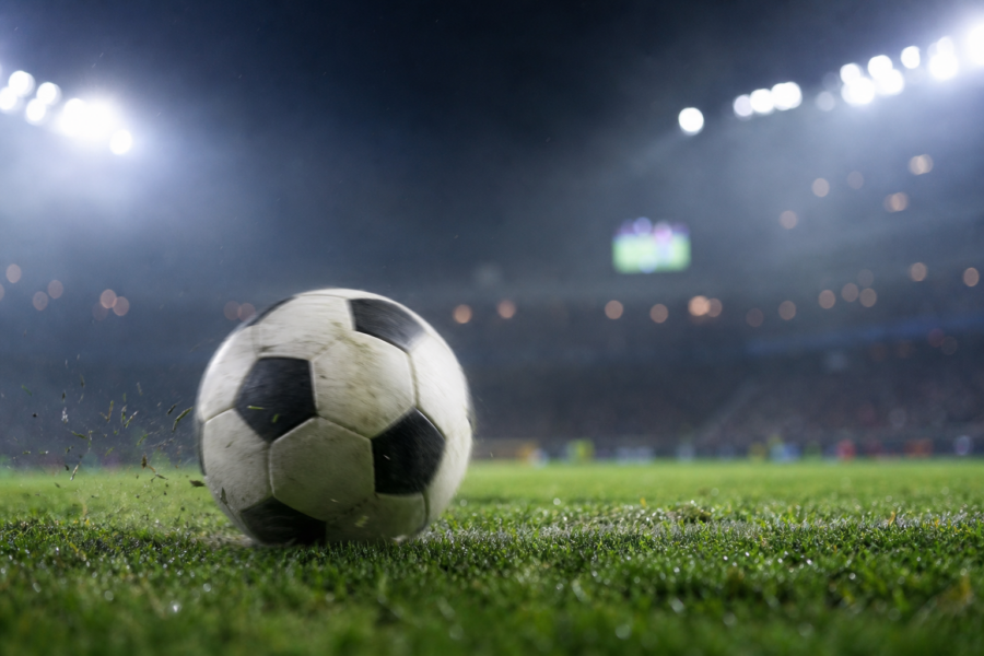 Bola de futebol genérica no gramado com estádio iluminado ao fundo, telão desfocado e clima de jogo.
