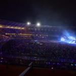 Arquibancadas iluminadas por lightsticks em noite de show no Brasil, com palco ao fundo.