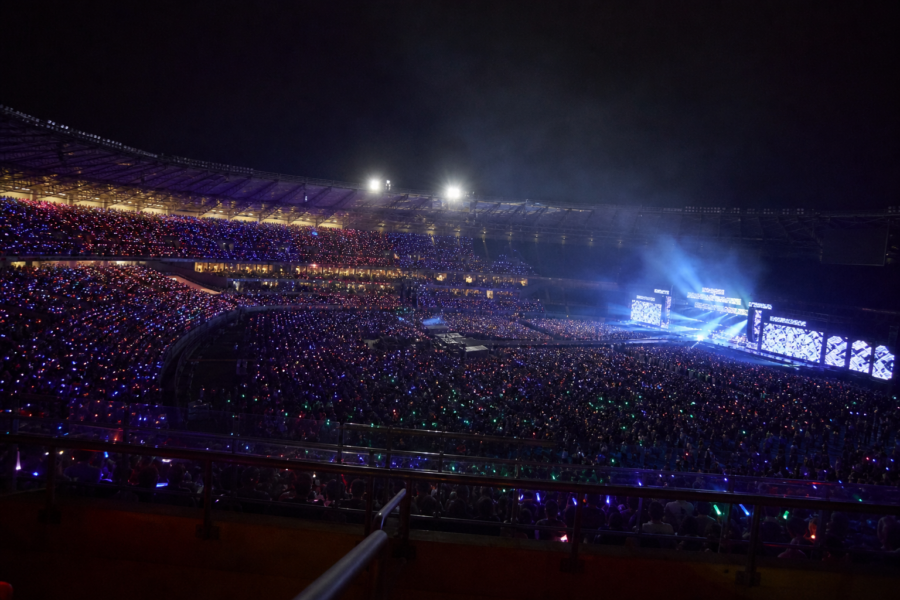 Arquibancadas iluminadas por lightsticks em noite de show no Brasil, com palco ao fundo.