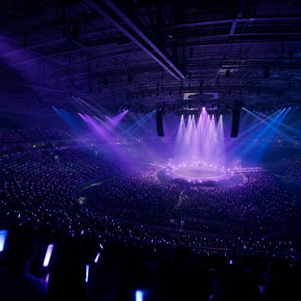 Arena lotada com luzes roxas, palco circular ao centro e público com bastões de luz durante uma estreia ao vivo.