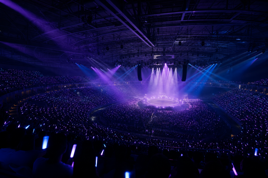 Arena lotada com luzes roxas, palco circular ao centro e público com bastões de luz durante uma estreia ao vivo.