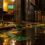Bandeirinhas do Brasil no chão molhado de uma rua após jogo, TV ao fundo desfocada com gramado.