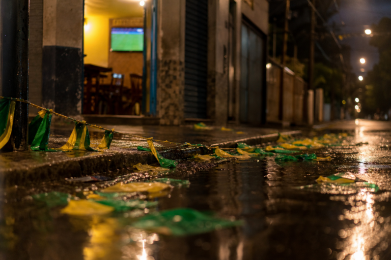 Bandeirinhas do Brasil no chão molhado de uma rua após jogo, TV ao fundo desfocada com gramado.