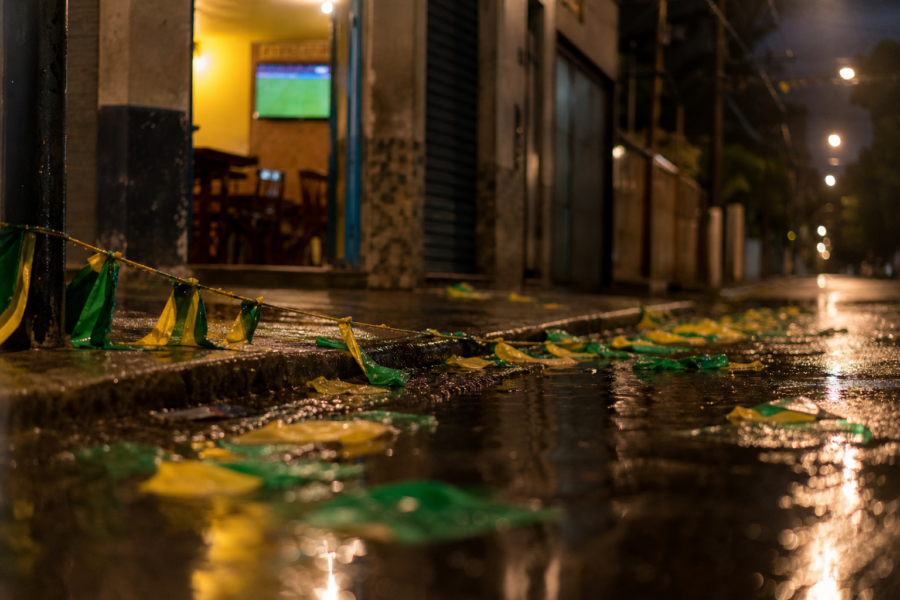Bandeirinhas do Brasil no chão molhado de uma rua após jogo, TV ao fundo desfocada com gramado.