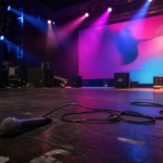 Bastidores de palco com luzes acesas e microfone no chão, prontos para show.