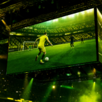 Telão de eSports com avatar de futebol e luzes verde e amarelas diante de arquibancada lotada, sem marcas visíveis.