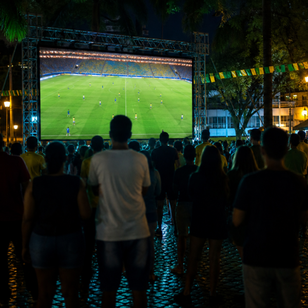 Telão em praça pública lotada transmitindo futebol ao vivo, torcida vista de longe e sem marcas visíveis.