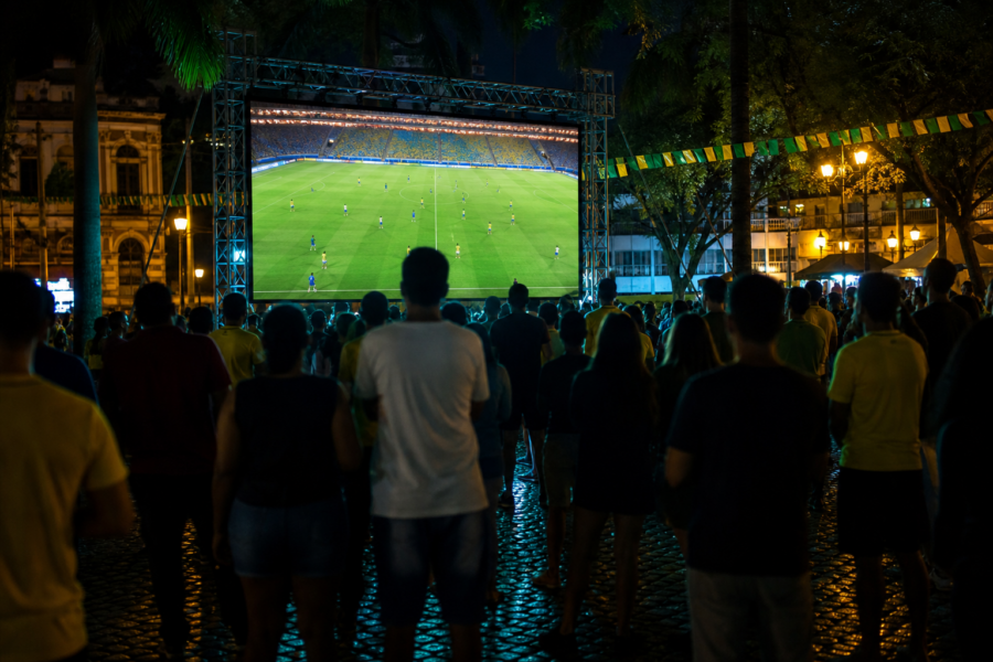 Telão em praça pública lotada transmitindo futebol ao vivo, torcida vista de longe e sem marcas visíveis.