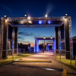 Entrada iluminada de um grande festival de música à noite, com estruturas e público ao fundo sem marcas visíveis.